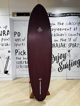 Maroon surfboard standing upright on a wooden floor inside a shop, with a wall covered in black text and drawings in the background. A small white logo is near the center.