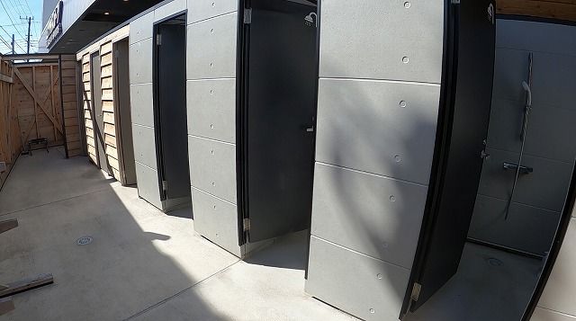 554d44e28c989f653c55f6afd74ded86 1 - SHOP NEWS | ムラサキスポーツオンラインストア 通販 Row of grey metal storage unit doors along a concrete alley, with a wooden fence on the left and sunlight casting shadows on the ground.