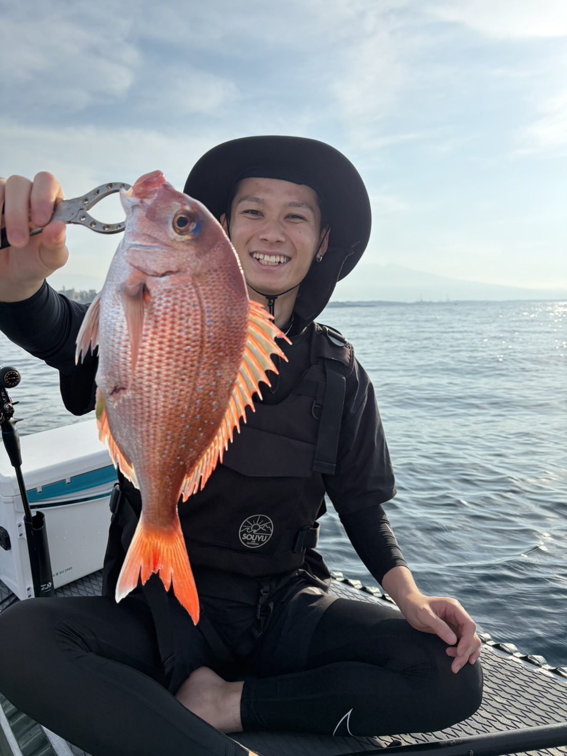 Smiling person in a wide-brimmed hat on a boat, holding a large red fish with a fish gripper against the sea and sky.