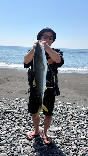 Person on a pebbly beach holding a large yellowtail-like fish up to chest, wearing a hat, sunglasses, black shirt and shorts.