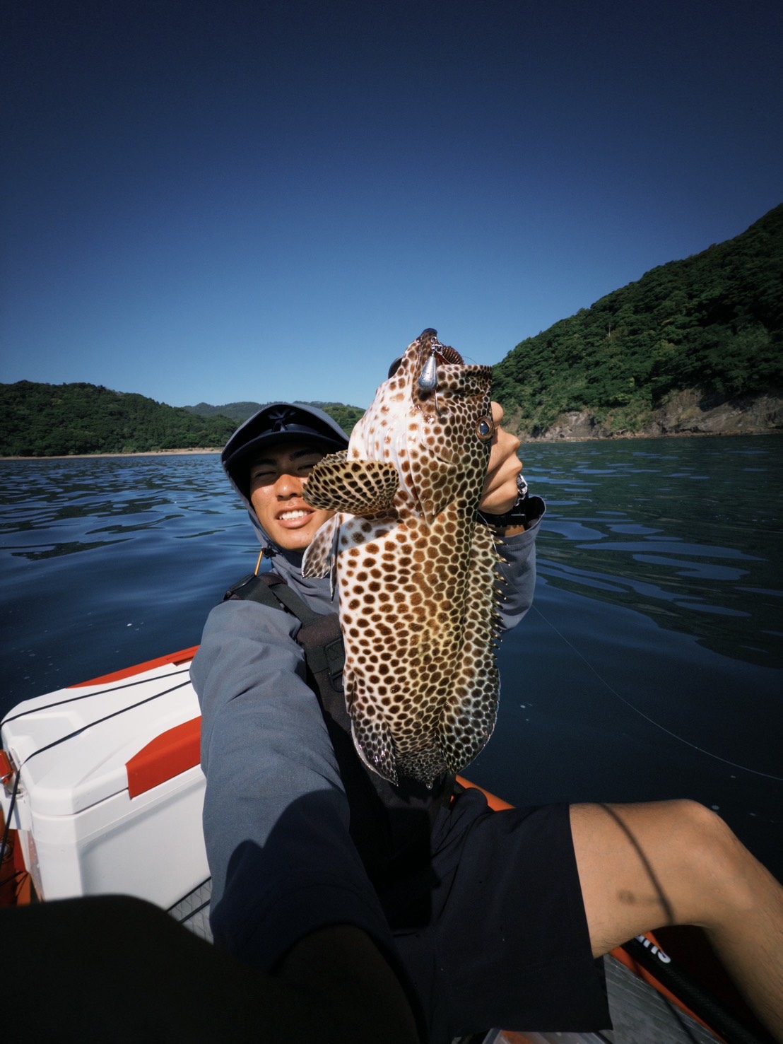 Person on a small boat holding up a large leopard-spotted fish against blue water and green hills in the background, smiling at the camera.