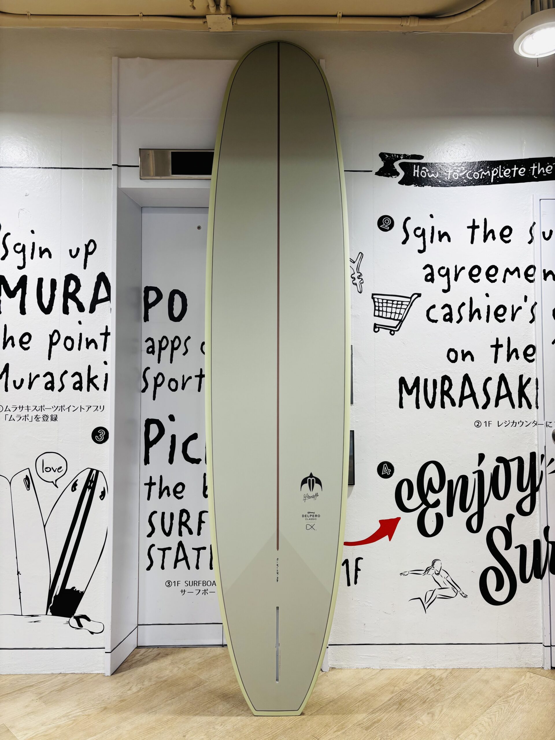 IMG_0239 - SHOP NEWS | ムラサキスポーツオンラインストア 通販 A pale-green surfboard stands upright against a wall covered with black handwritten surfing text and drawings in a gallery-like space.