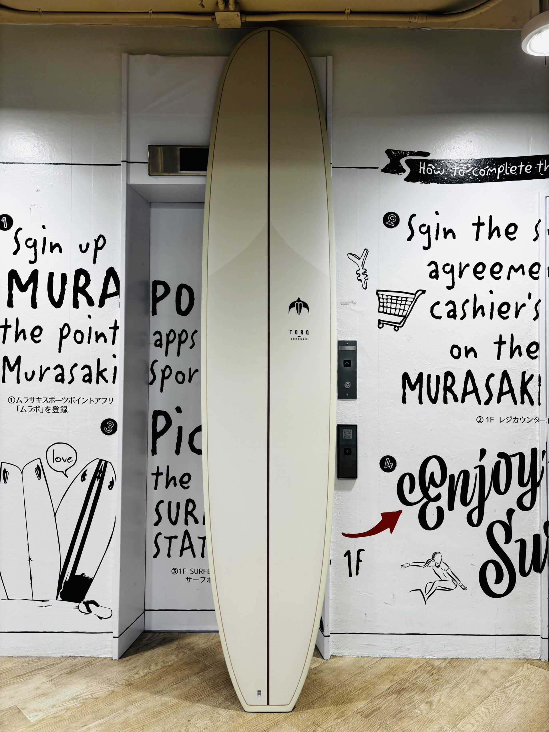 IMG_0240 - SHOP NEWS | ムラサキスポーツオンラインストア 通販 Beige surfboard standing upright between graffiti-covered walls with black handwritten text, indoors on a wooden floor.