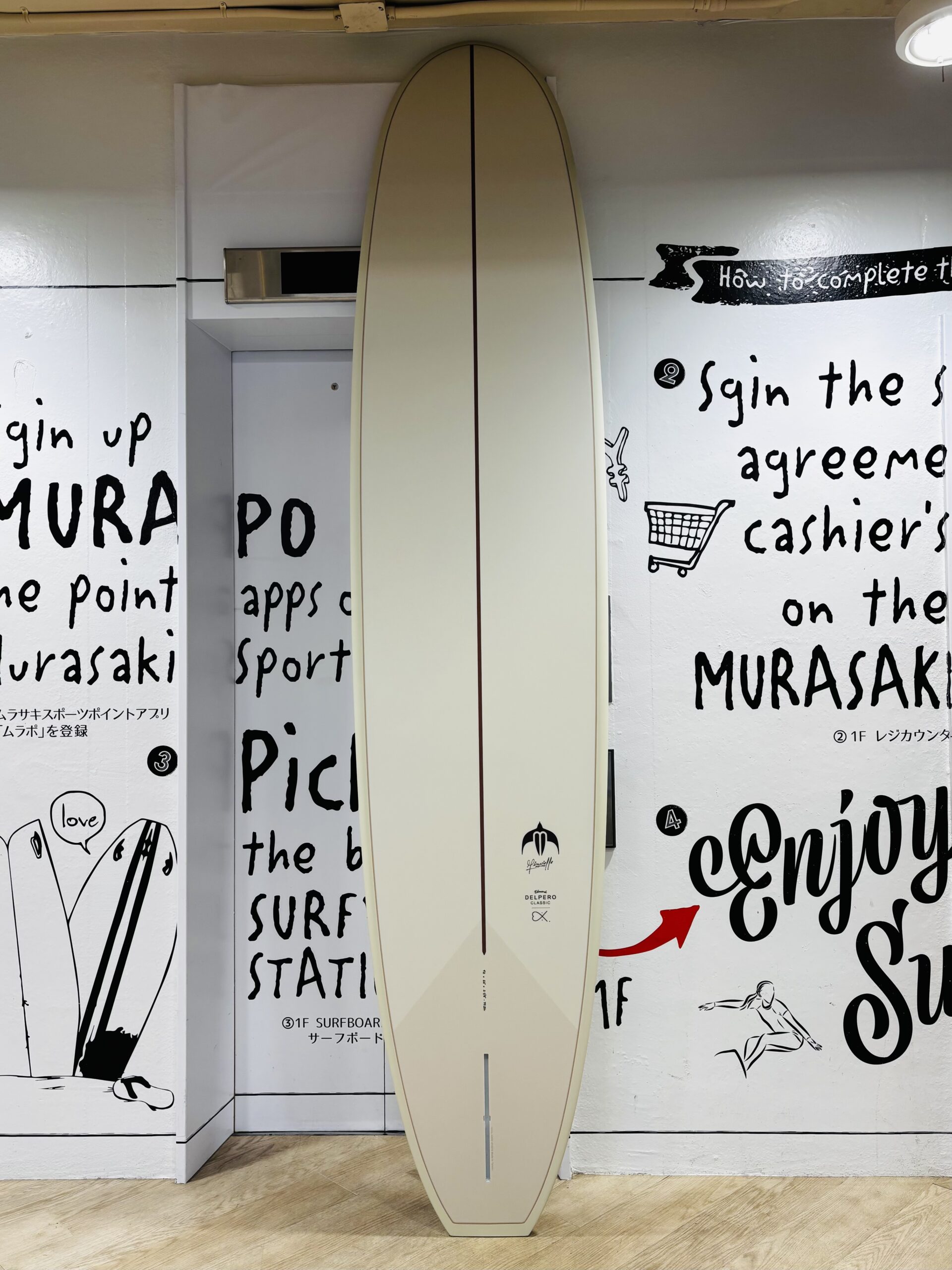 IMG_0241 - SHOP NEWS | ムラサキスポーツオンラインストア 通販 Pale beige surfboard standing upright in a showroom between white walls with bold black handwritten text and small drawings on them.