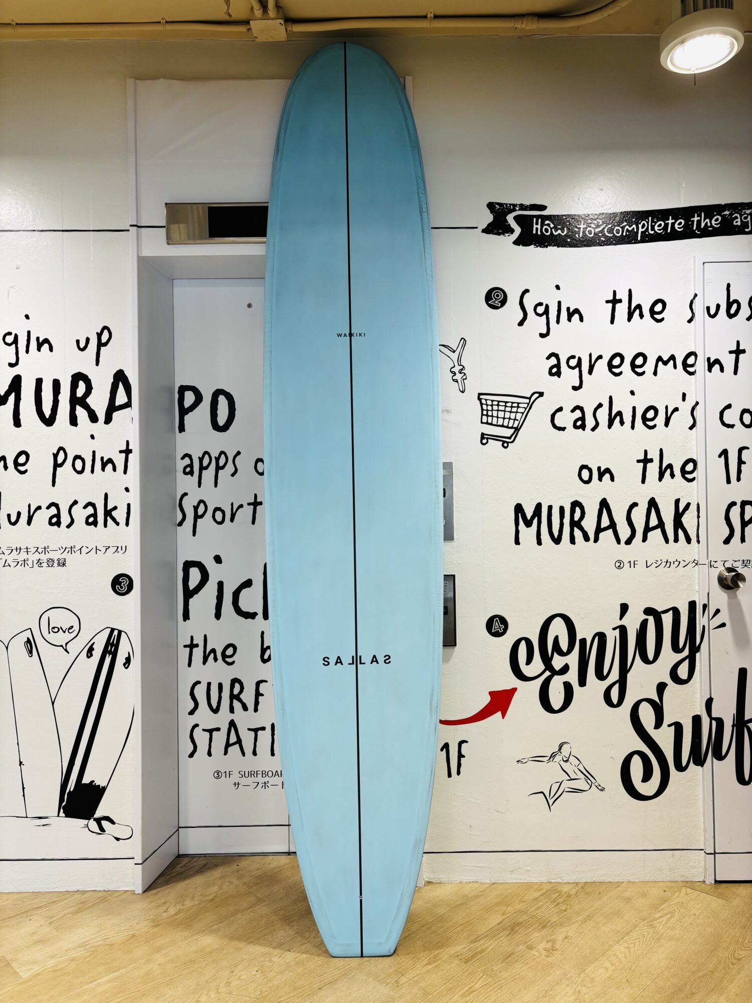 Blue surfboard leans upright against a white wall covered with black handwritten text and drawings, in a room with a wooden floor.