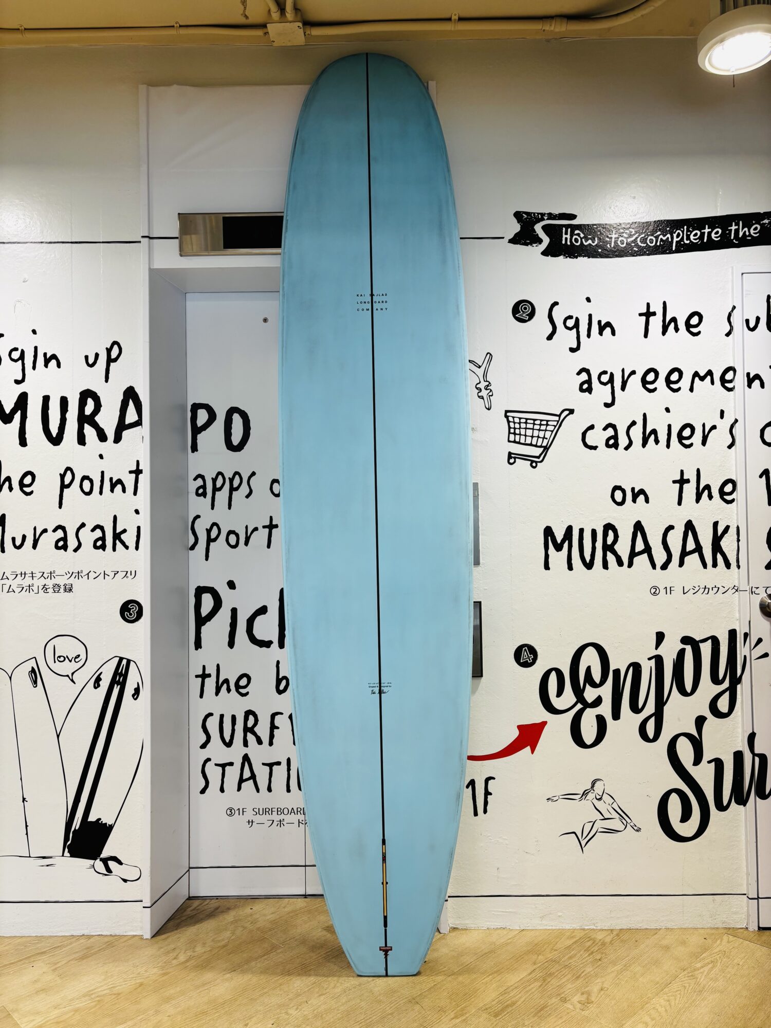 Blue surfboard stands upright against a white wall covered in black handwritten text and small drawings in a shop interior.