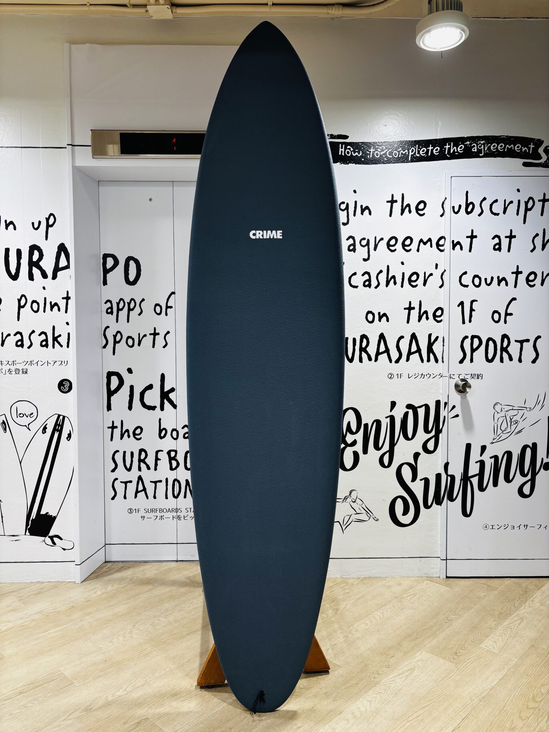 Blue surfboard standing upright indoors in a showroom, with surfing-themed wall text in the background