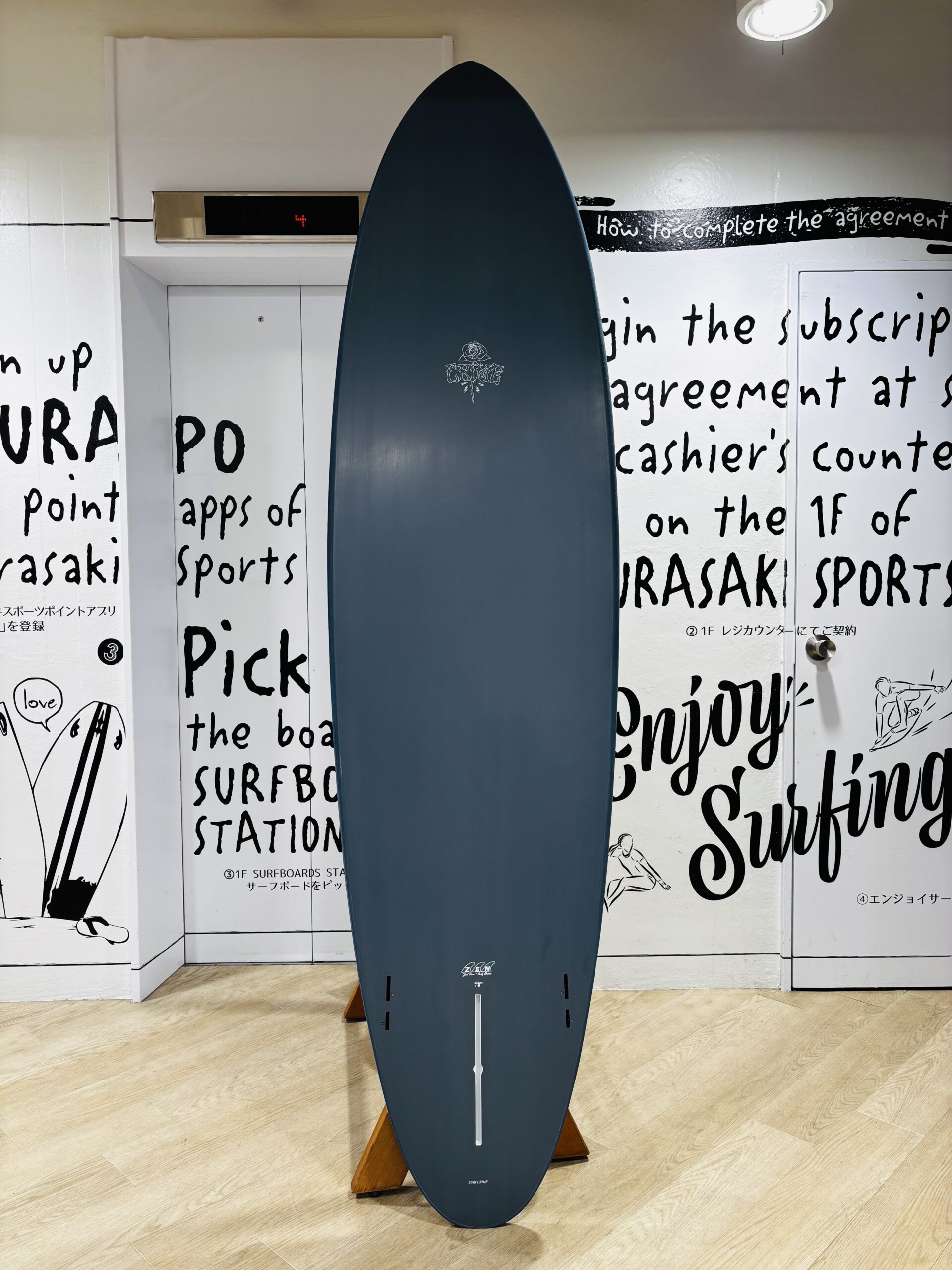 Tall blue surfboard on a wooden stand indoors, positioned in front of a wall with black handwritten text art on white panels.