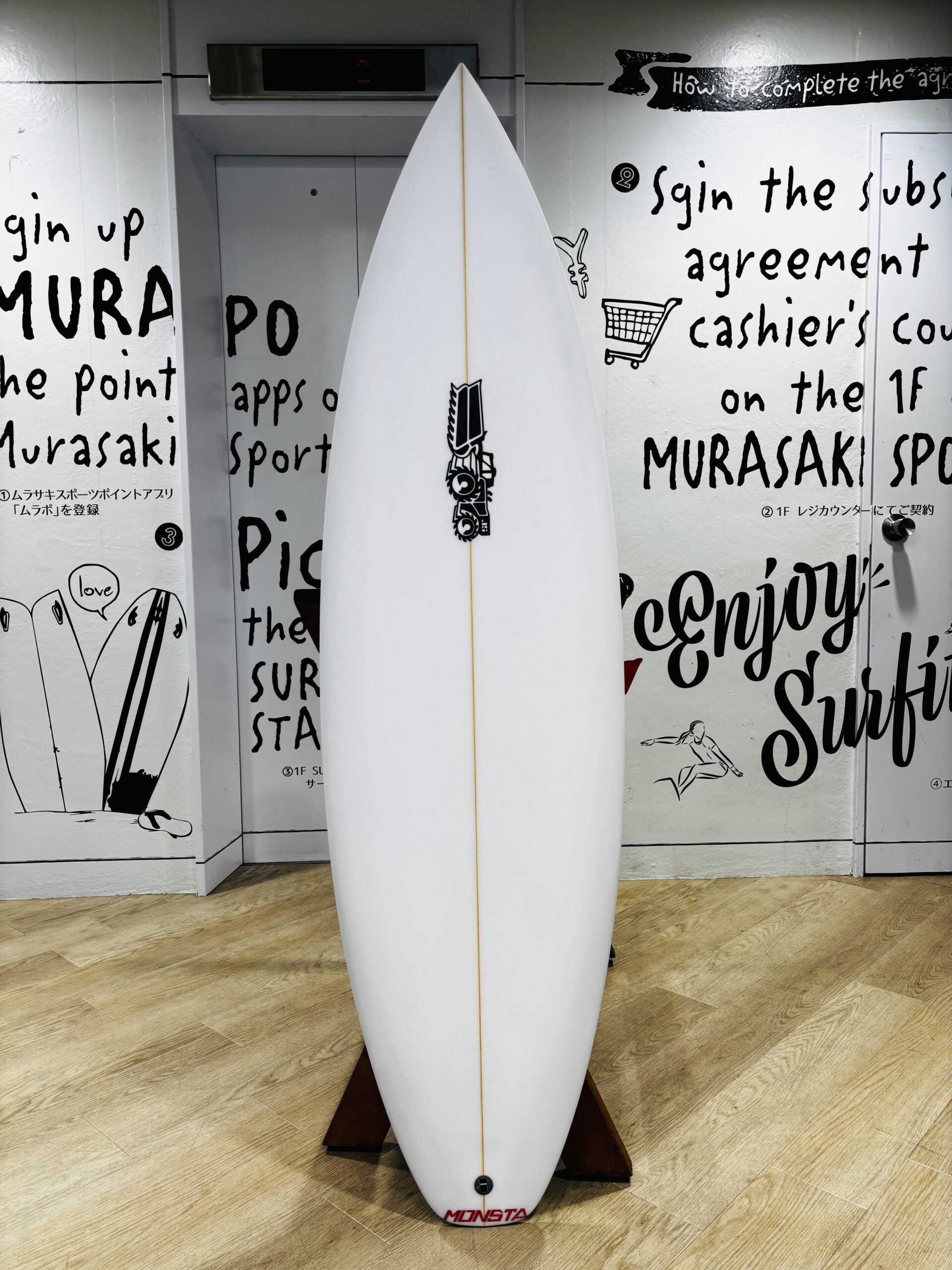 IMG_0789 - SHOP NEWS | ムラサキスポーツオンラインストア 通販 White surfboard on a wooden stand in a room with graffiti-style black text on white walls and a light wood floor; MONSTA logo in red near the tail.