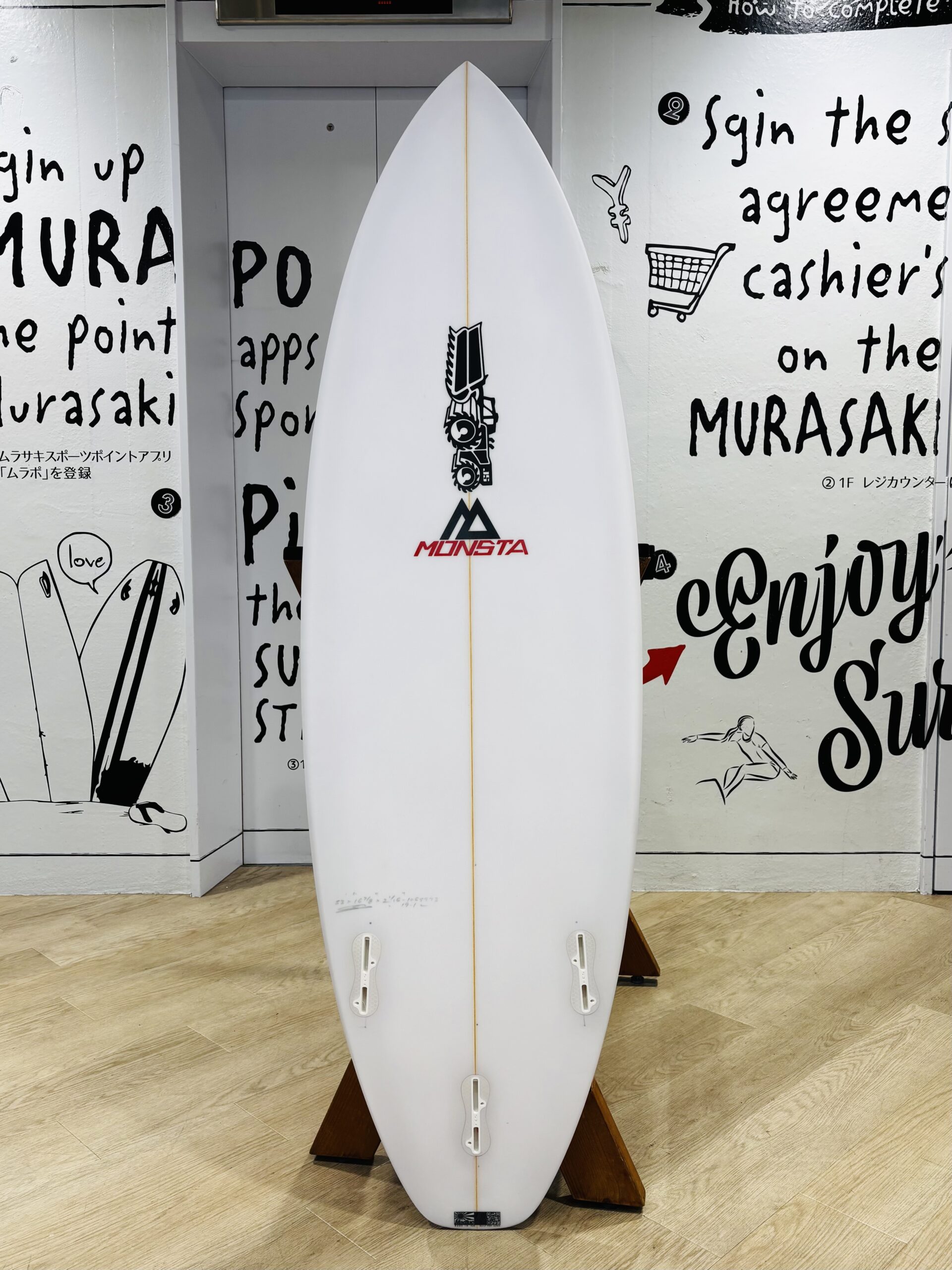IMG_0790 - SHOP NEWS | ムラサキスポーツオンラインストア 通販 White surfboard on a wooden stand indoors, displaying the MONSTA logo against a graffiti-covered wall.