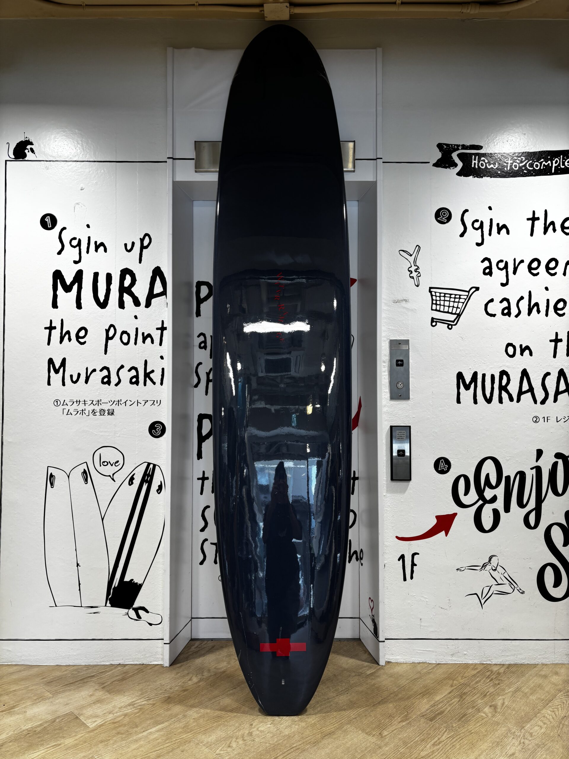 Tall glossy black surfboard standing upright in a white-walled display alcove with graffiti-style text and drawings on either side, and a wooden floor beneath.