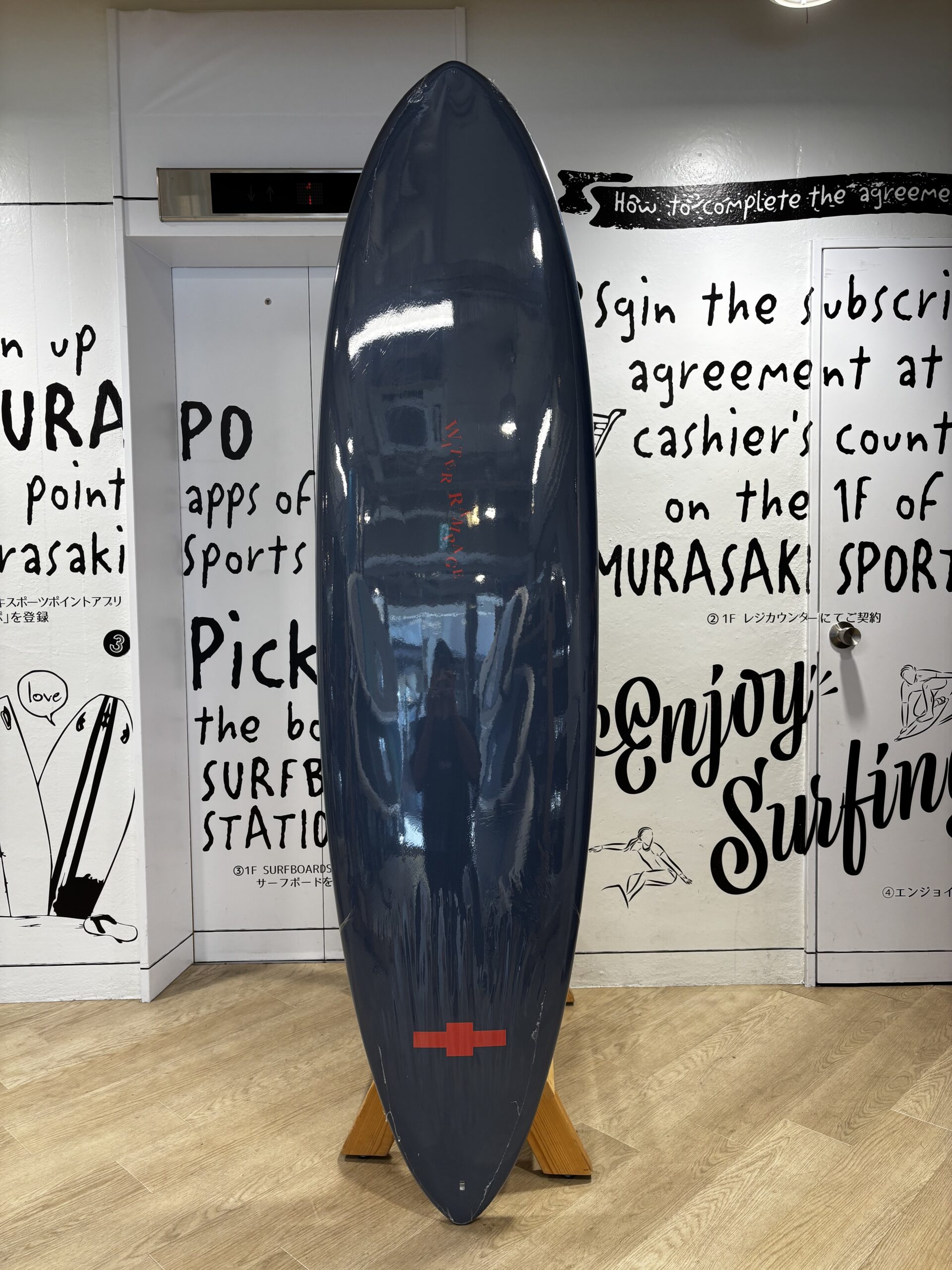 Blue surfboard standing upright on a wooden support indoors, with black handwritten wall text in the background and a red cross near the bottom.