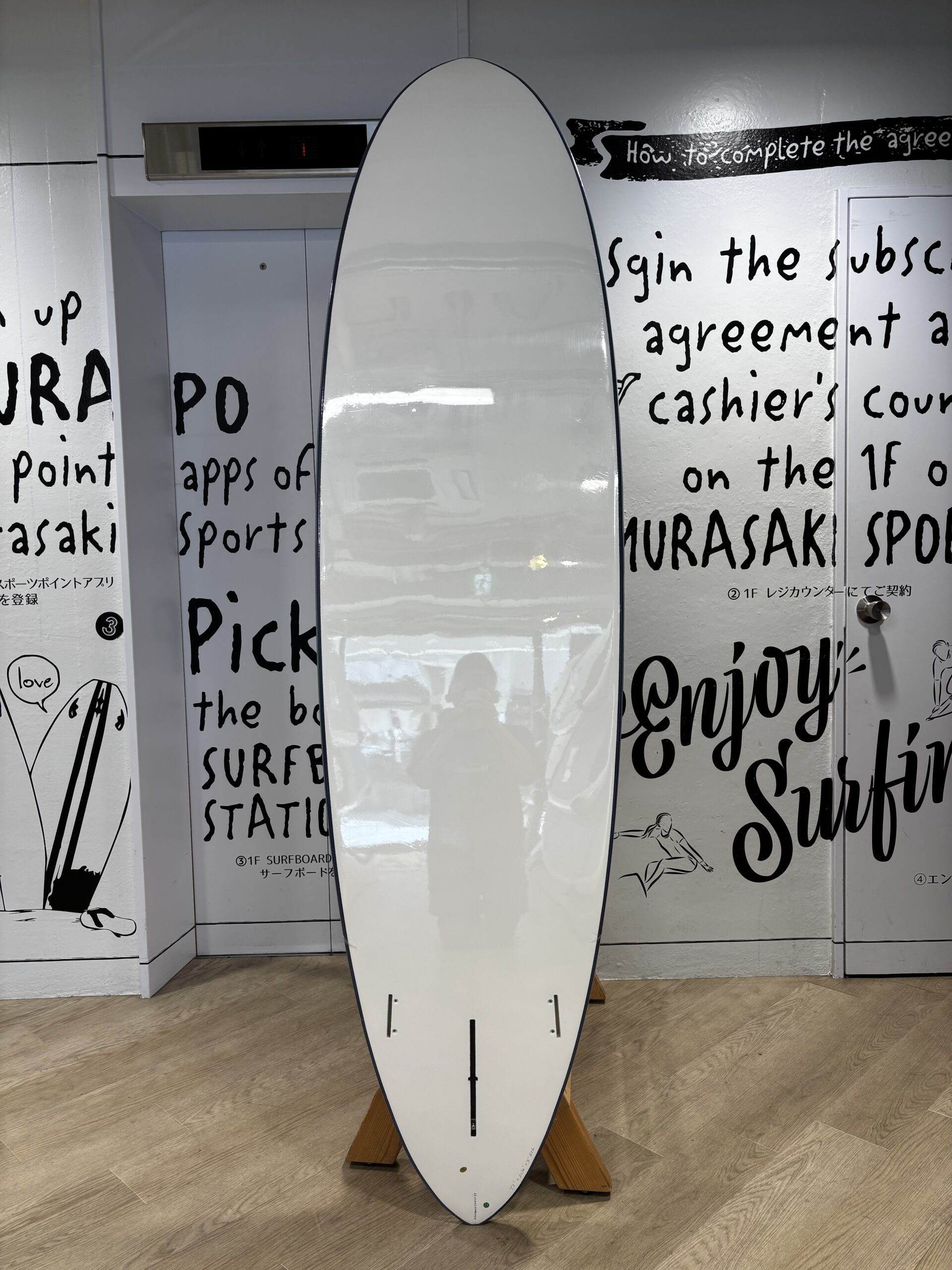 White surfboard on a wooden stand indoors, with a wall of black handwritten text and simple drawings behind it.