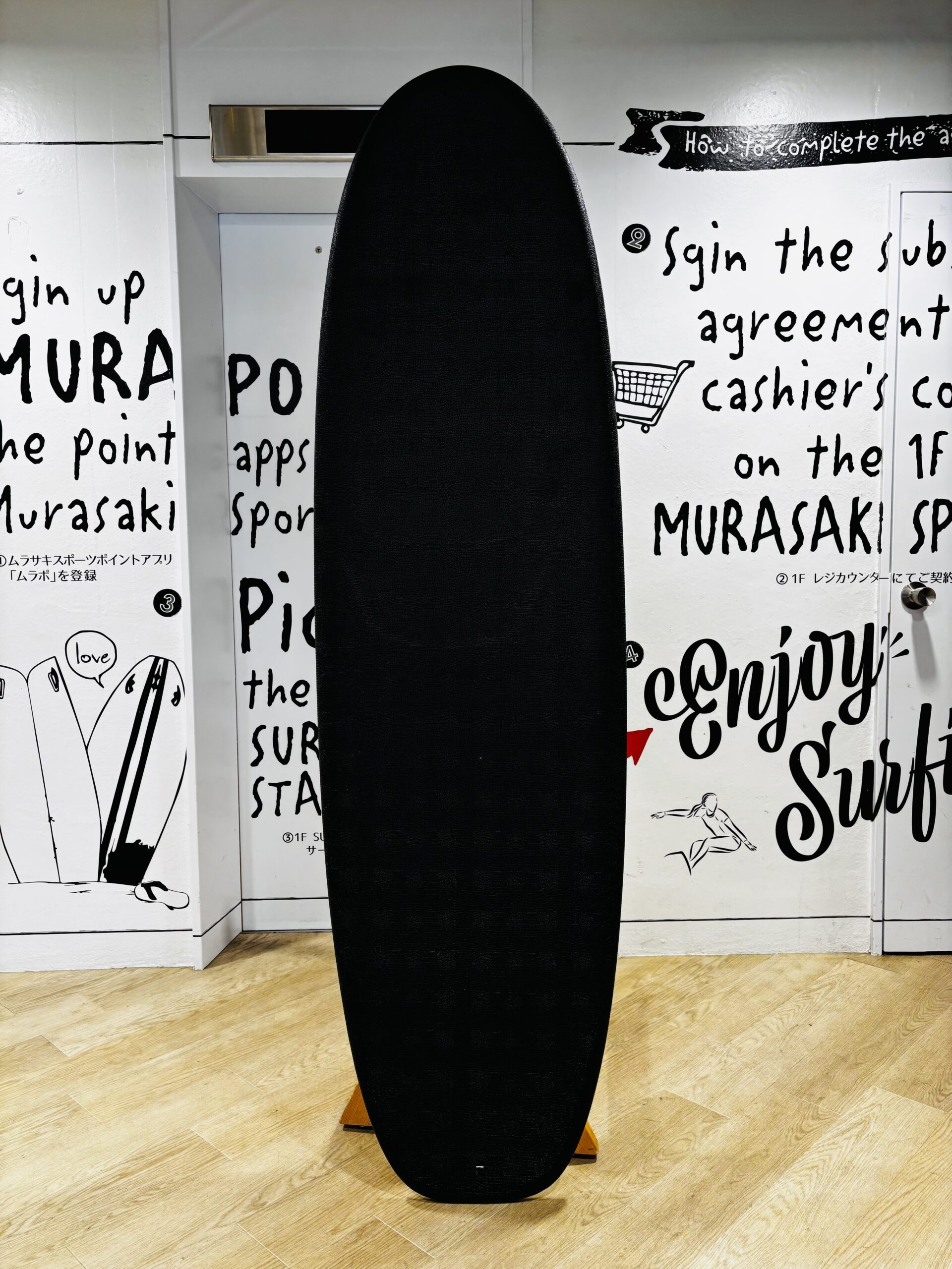 Vertical black surfboard-shaped object standing upright in a room with white walls covered in handwritten text and drawings, on a wooden floor.