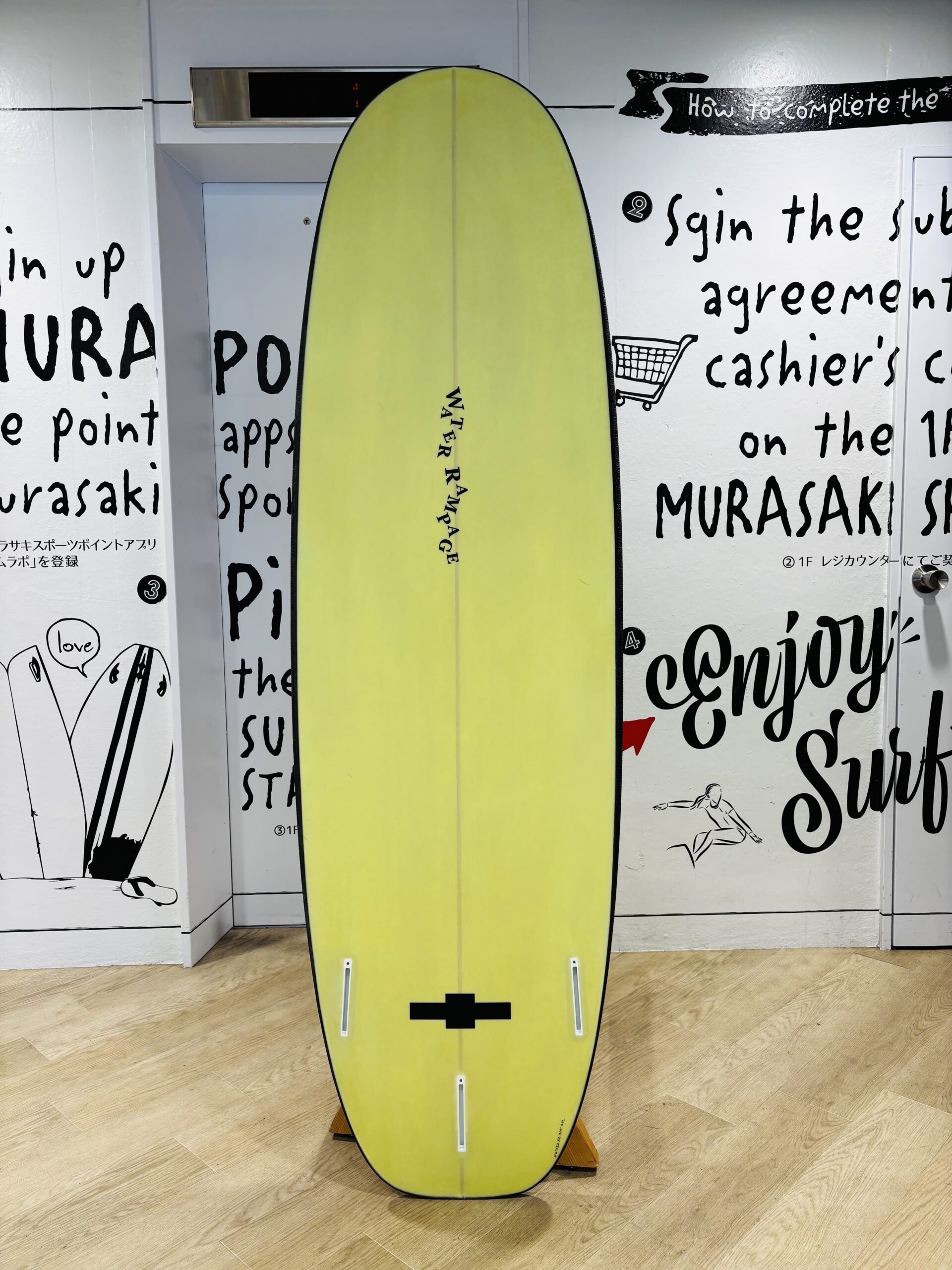 Yellow surfboard displayed indoors on a wooden floor, with bold black text graphics on white walls behind the board.