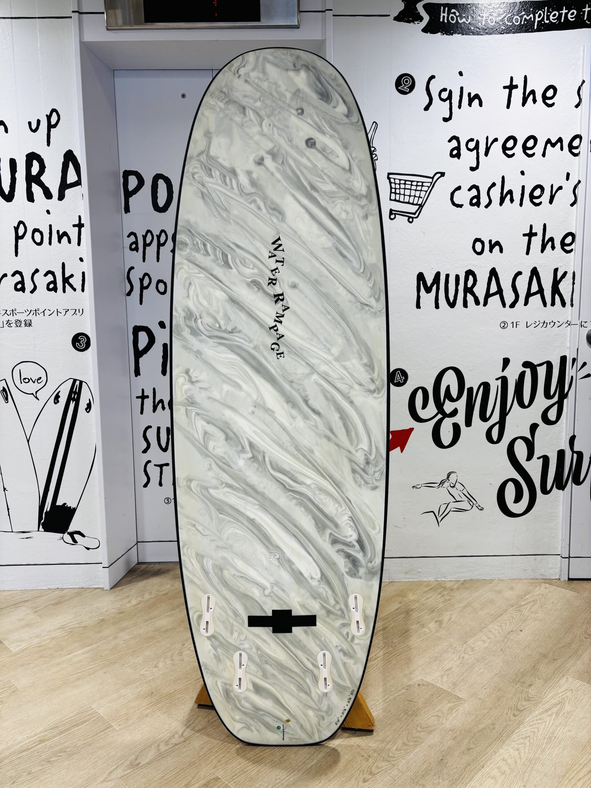 Marble-gray surfboard upright on a wooden stand inside a white-walled room with large black handwritten text on the walls; board has multiple fin boxes and 'WATER RAMPAGE' written along its center.