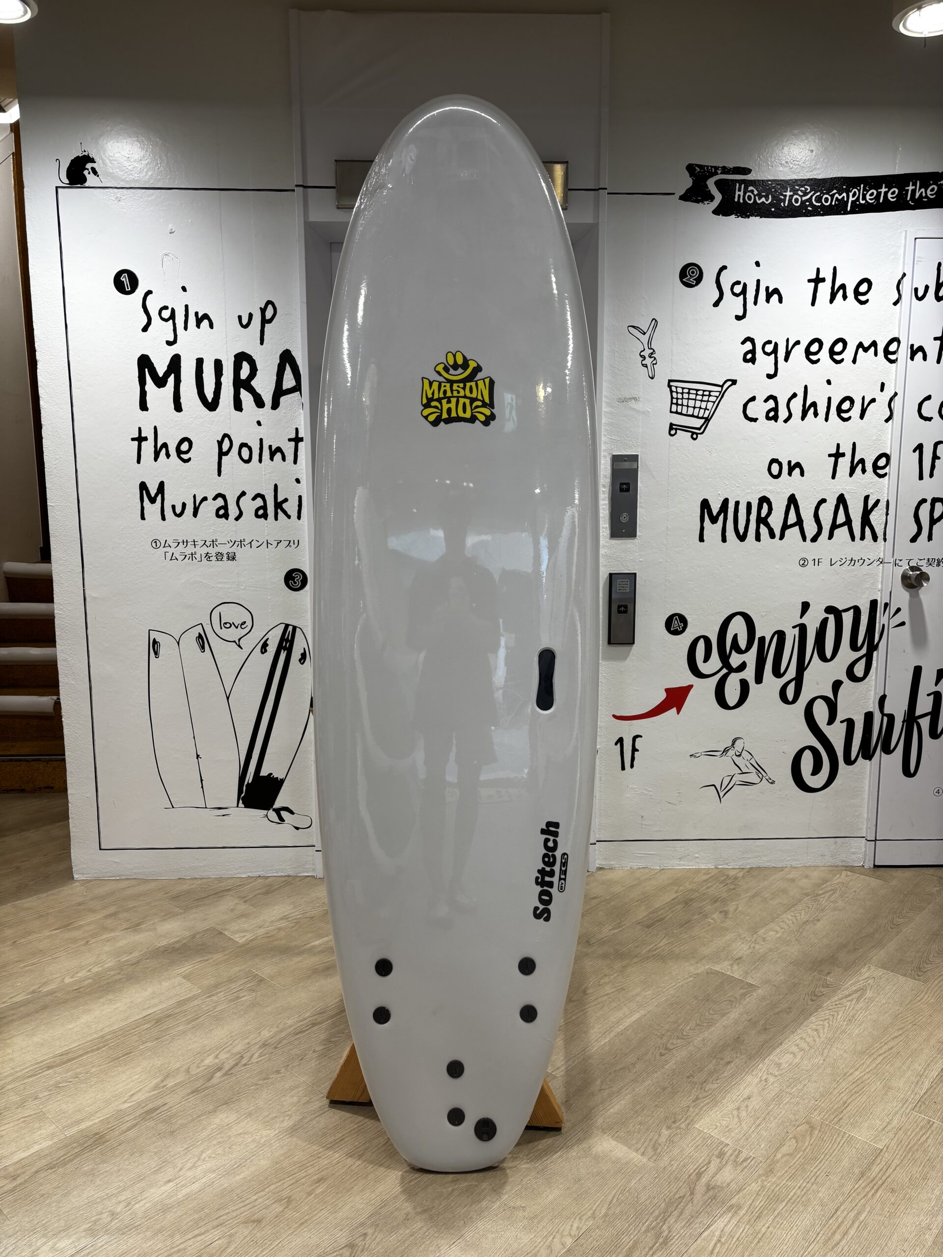 Gray surfboard on a wooden stand indoors, with black graffiti-style wall art behind it in a showroom setting.