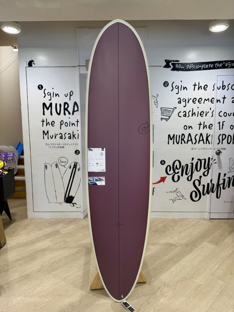 IMG_5061 - SHOP NEWS | ムラサキスポーツオンラインストア 通販 Purple longboard surfboard standing vertically on a wooden rack inside a shop, with surf-themed posters on the walls behind it.