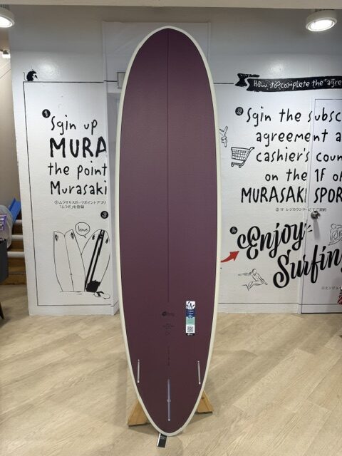 IMG_5062 - SHOP NEWS | ムラサキスポーツオンラインストア 通販 Purple surfboard on a wooden stand inside a shop, with graffiti-style wall art on doors in the background.