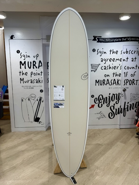 IMG_5064 - SHOP NEWS | ムラサキスポーツオンラインストア 通販 Cream-colored surfboard on a wooden rack standing upright inside a shop, with wall graphics and handwritten signs in the background.
