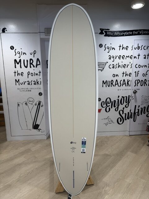 IMG_5065 - SHOP NEWS | ムラサキスポーツオンラインストア 通販 Cream-colored surfboard with a subtle orange stringer, standing upright on a wooden display rack inside a shop with graphic walls.