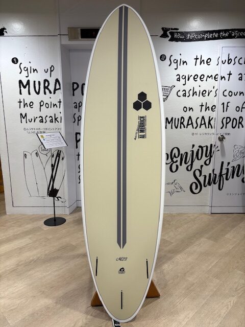 IMG_7070 - SHOP NEWS | ムラサキスポーツオンラインストア 通販 Cream surfboard with two dark grey stripes, upright on a stand in a shop, featuring a small hexagonal logo.