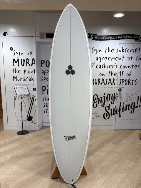 IMG_7071 - SHOP NEWS | ムラサキスポーツオンラインストア 通販 White surfboard on a wooden stand indoors, centered, with a small hexagonal logo along its stringer; graffiti-style wall and signage in the background.