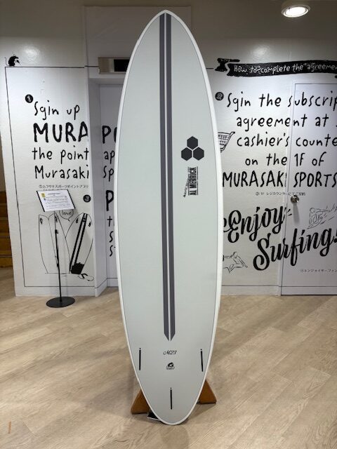 IMG_7072 - SHOP NEWS | ムラサキスポーツオンラインストア 通販 White surfboard on a wooden stand in a shop, featuring a dark gray center stripe and logo, with 'Enjoy Surfing' wall art in the background.