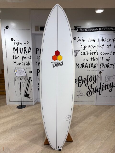 IMG_7073 - SHOP NEWS | ムラサキスポーツオンラインストア 通販 White surfboard with a red and yellow hexagonal logo centered, displayed upright on a wooden stand inside a surf shop with black graffiti-style wall art.