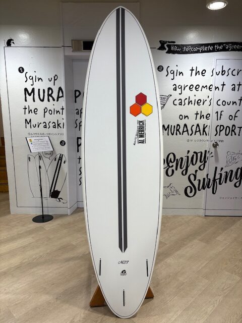 IMG_7074 - SHOP NEWS | ムラサキスポーツオンラインストア 通販 White surfboard with two dark gray center stripes stands on a wooden rack in a store; a hexagon logo and vertical label on the right side, with wall graphics and display stands in the background.