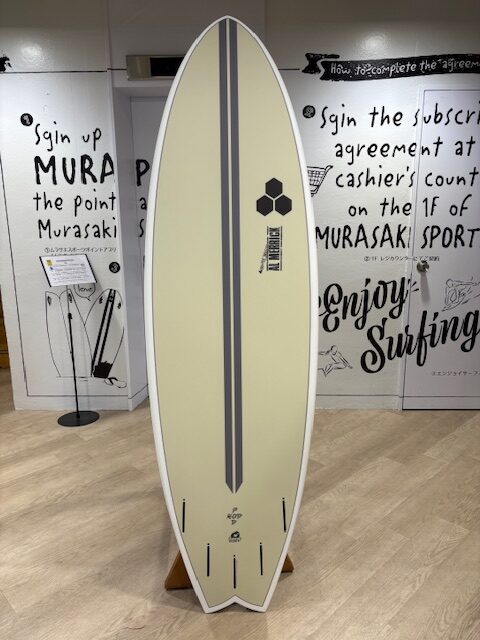 IMG_7076 - SHOP NEWS | ムラサキスポーツオンラインストア 通販 Cream surfboard with two dark gray stripes and a hexagonal logo, standing upright on a wooden rack in a surf shop showroom.