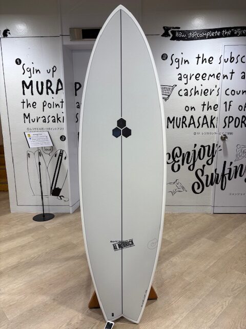 IMG_7077 - SHOP NEWS | ムラサキスポーツオンラインストア 通販 White surfboard on a stand indoors, featuring a vertical stringer, a small black hexagonal logo midboard, and a brand sticker near the tail.