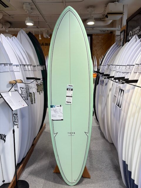 IMG_7380 - SHOP NEWS | ムラサキスポーツオンラインストア 通販 Mint-green surfboard standing upright on a showroom display, with price tags attached and rows of white boards on either side in a shop.