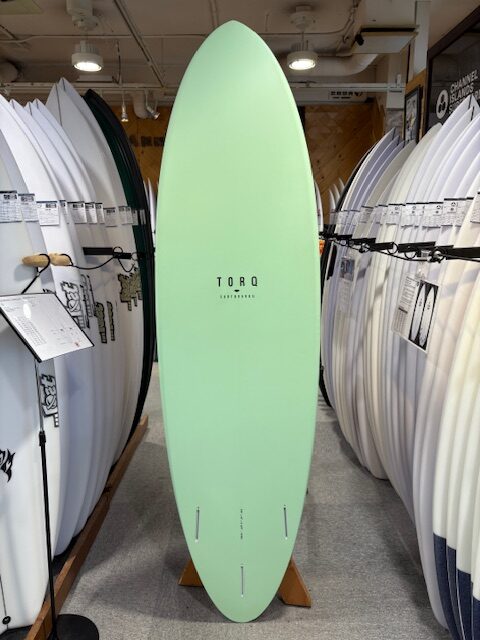 IMG_7381 - SHOP NEWS | ムラサキスポーツオンラインストア 通販 Mint green longboard surfboard on a display stand in a surf shop, surrounded by white boards on racks, center brand reads TORO Surfboards