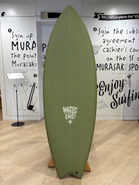 IMG_8793 - SHOP NEWS | ムラサキスポーツオンラインストア 通販 Green surfboard on a wooden stand in a showroom with black graffiti-style wall text behind it; has a white 'Magic Surf' logo near the bottom center of the board