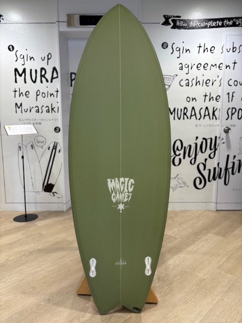IMG_8794 - SHOP NEWS | ムラサキスポーツオンラインストア 通販 Olive-green surfboard on a wooden stand inside a showroom, featuring a white 'Magic Carpet' logo and white fin boxes at the base.
