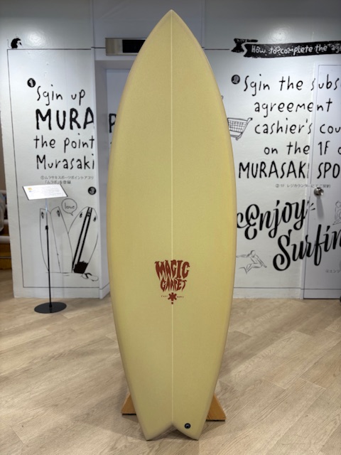 IMG_8798 - SHOP NEWS | ムラサキスポーツオンラインストア 通販 Cream-colored surfboard on a wooden stand, displayed vertically in a gallery with a 'Magic Carpet' logo in red at center.