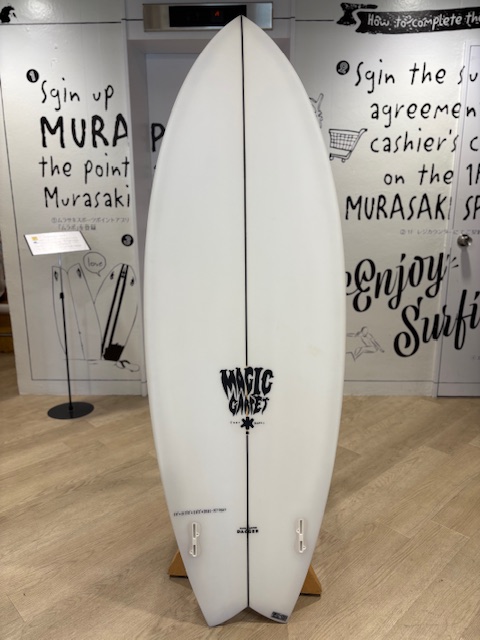 IMG_8802 - SHOP NEWS | ムラサキスポーツオンラインストア 通販 White surfboard with a black center stringer on a wooden display rack in a surf shop, walls covered with surfing-themed typography.
