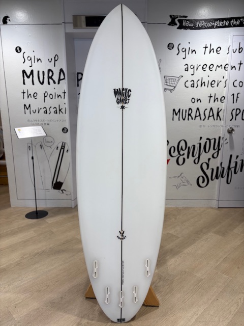 IMG_8809 - SHOP NEWS | ムラサキスポーツオンラインストア 通販 White surfboard on a wooden stand in a shop display, featuring a 'Magic Carpet' logo along its center.