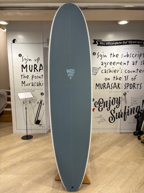 IMG_8819 - SHOP NEWS | ムラサキスポーツオンラインストア 通販 Blue-gray surfboard on a wooden stand in a shop, with black-and-white wall art behind.