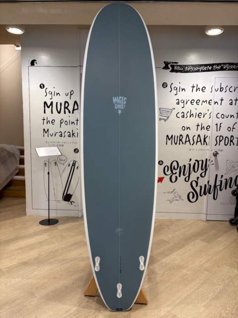IMG_8820 - SHOP NEWS | ムラサキスポーツオンラインストア 通販 Blue-gray surfboard on a wooden stand displayed indoors for sale, with white rails and small fins at the bottom and surfing-themed artwork on the walls behind.