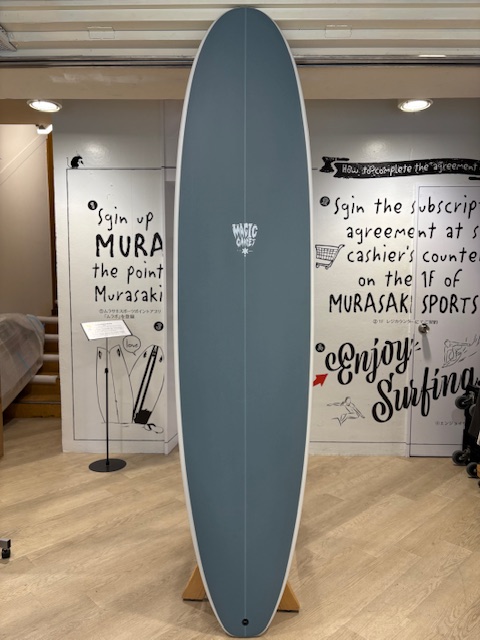 IMG_8823 - SHOP NEWS | ムラサキスポーツオンラインストア 通販 Blue-gray surfboard with a white edge, standing upright on a wooden display rack inside a shop showroom.