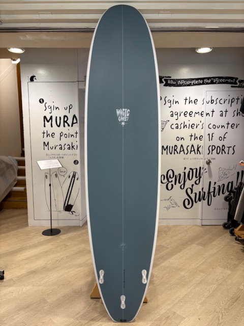 IMG_8826 - SHOP NEWS | ムラサキスポーツオンラインストア 通販 Blue/gray longboard surfboard with white edges standing upright on a display rack in a shop, with black-and-white wall graphics and wooden floor behind it.