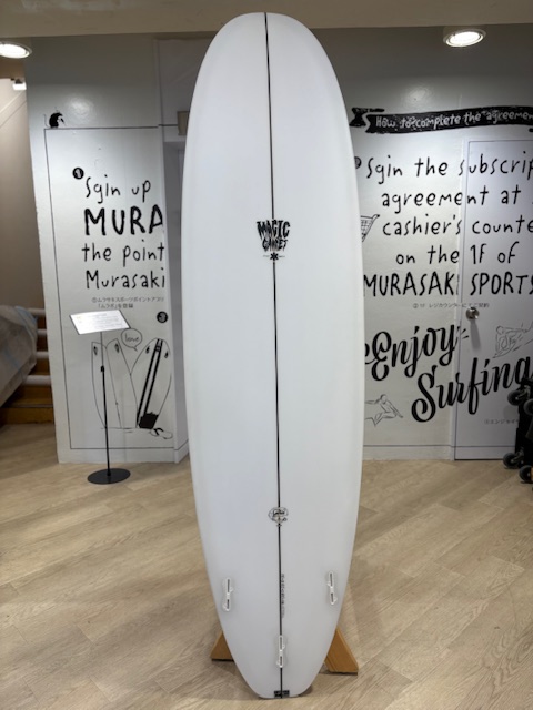 IMG_8829 - SHOP NEWS | ムラサキスポーツオンラインストア 通販 White longboard surfboard on a display stand in a surf shop showroom, centered with a vertical stringer and small logos along the deck.