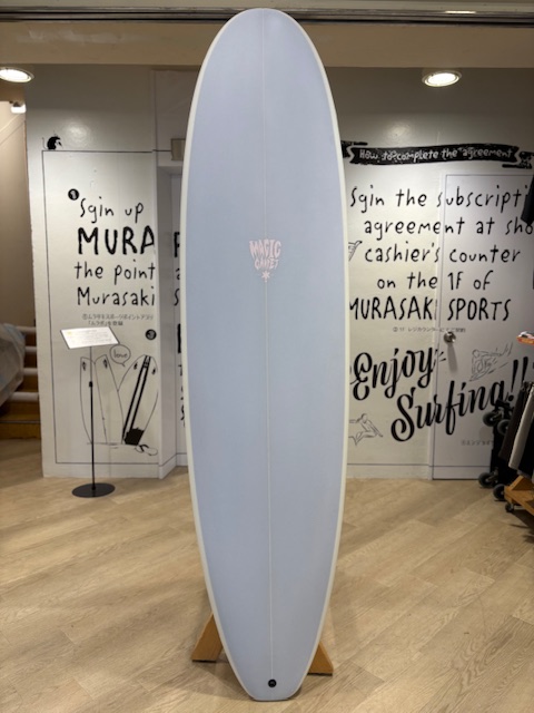 IMG_8830 - SHOP NEWS | ムラサキスポーツオンラインストア 通販 Light blue longboard surfboard on a wooden display stand in a shop, with handwritten wall art in the background.