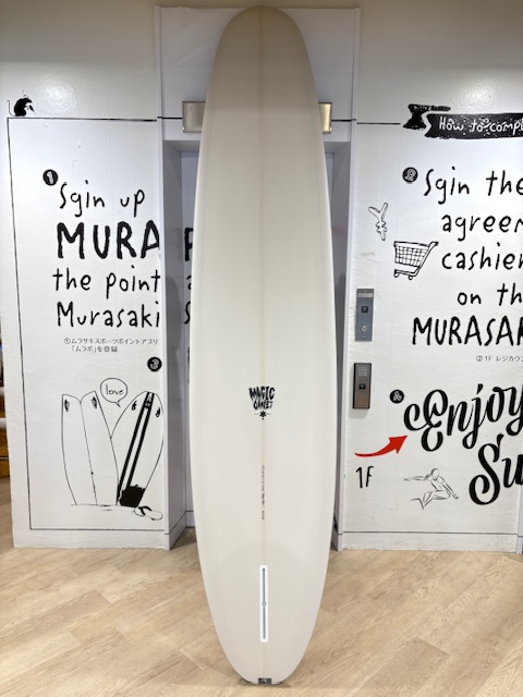 IMG_8849 - SHOP NEWS | ムラサキスポーツオンラインストア 通販 White surfboard standing upright indoors between graffiti-covered walls in a shop, with a small logo near the center.