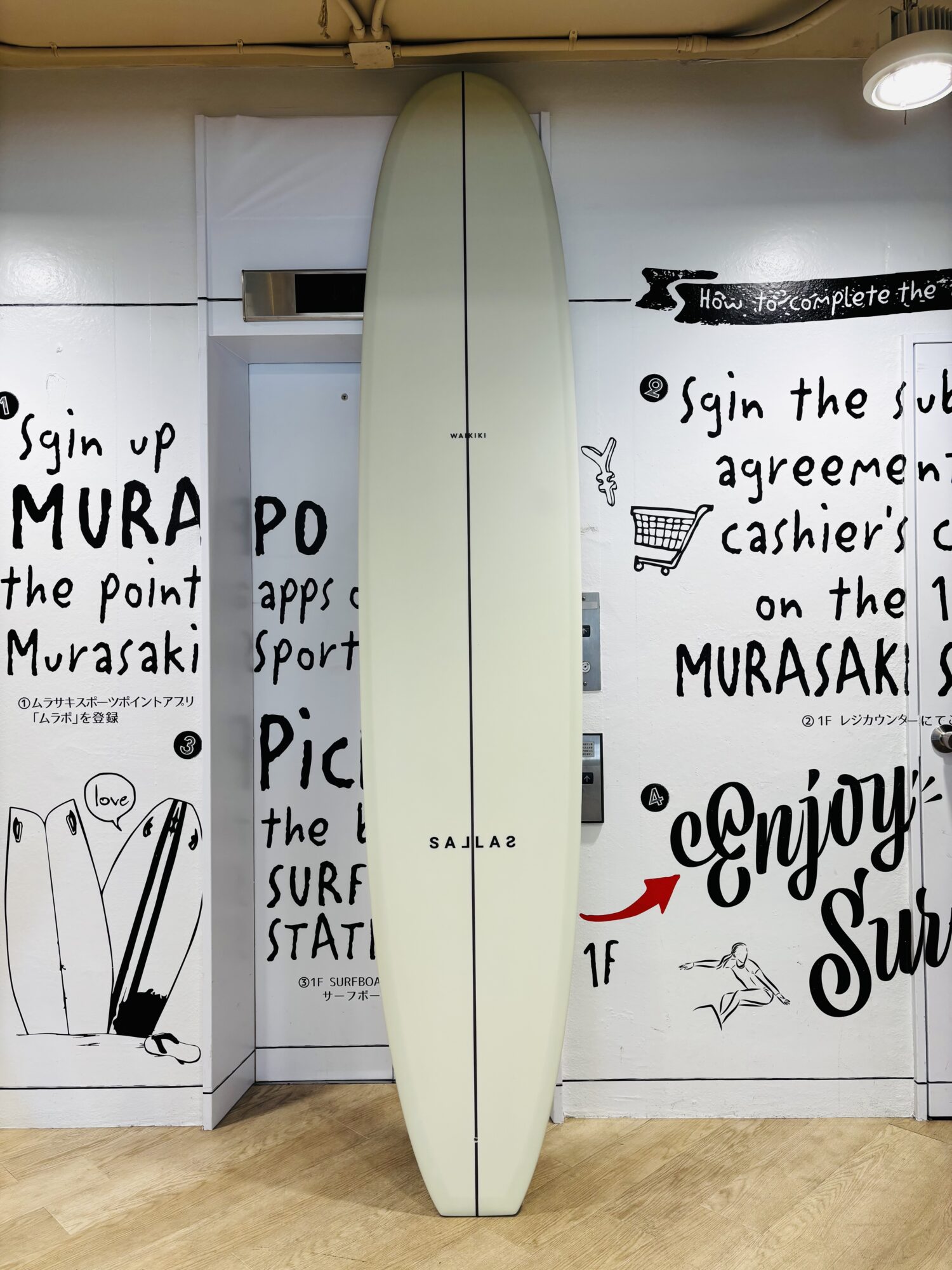 Off-white surfboard standing upright indoors against a wall with black graffiti-style text and illustrations.