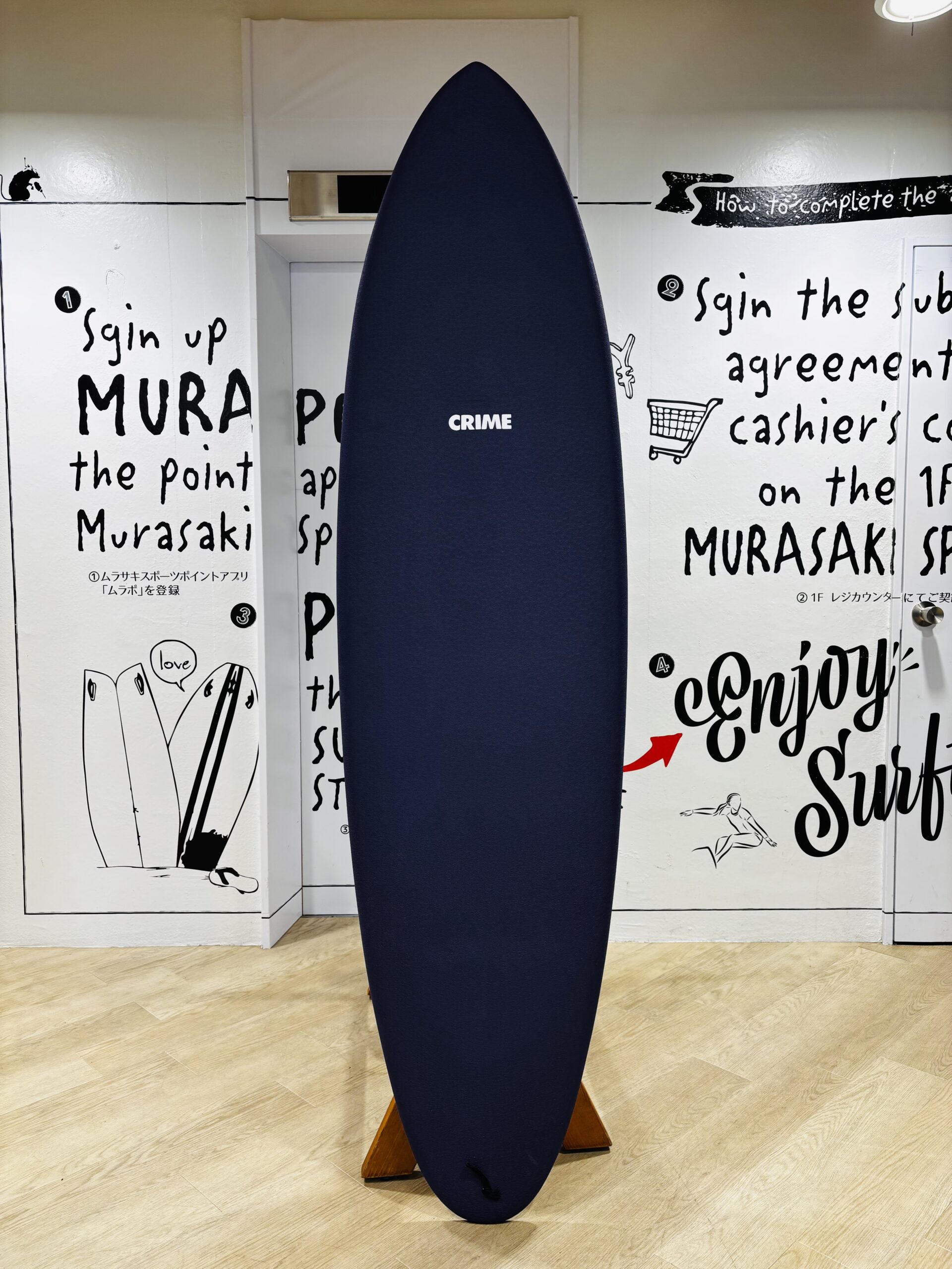Navy blue surfboard with a CRIME logo, standing upright on a wooden stand in a room with graffiti on white walls.