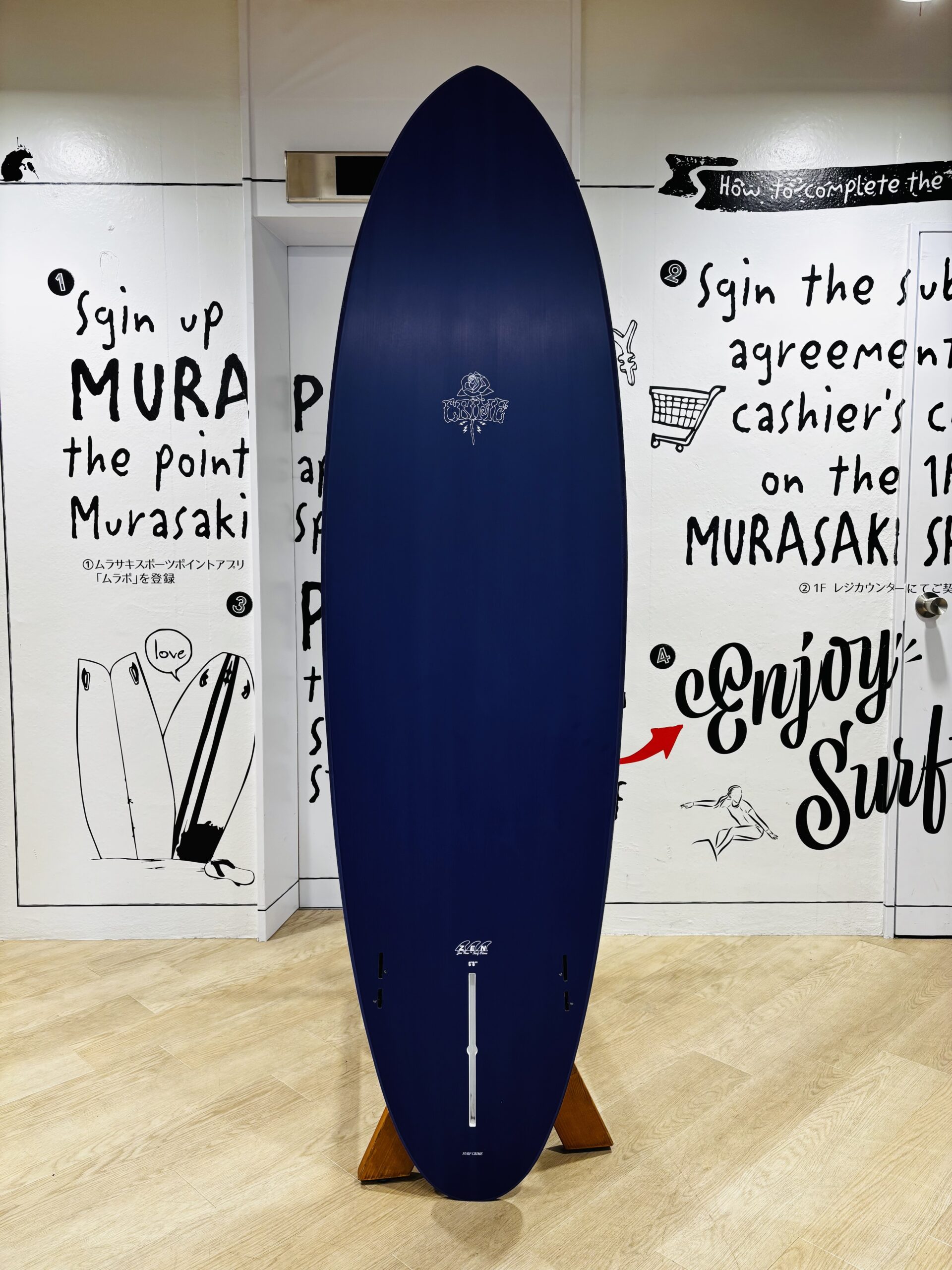 Blue surfboard on a wooden stand indoors, against wall art with surfing-themed text and drawings.