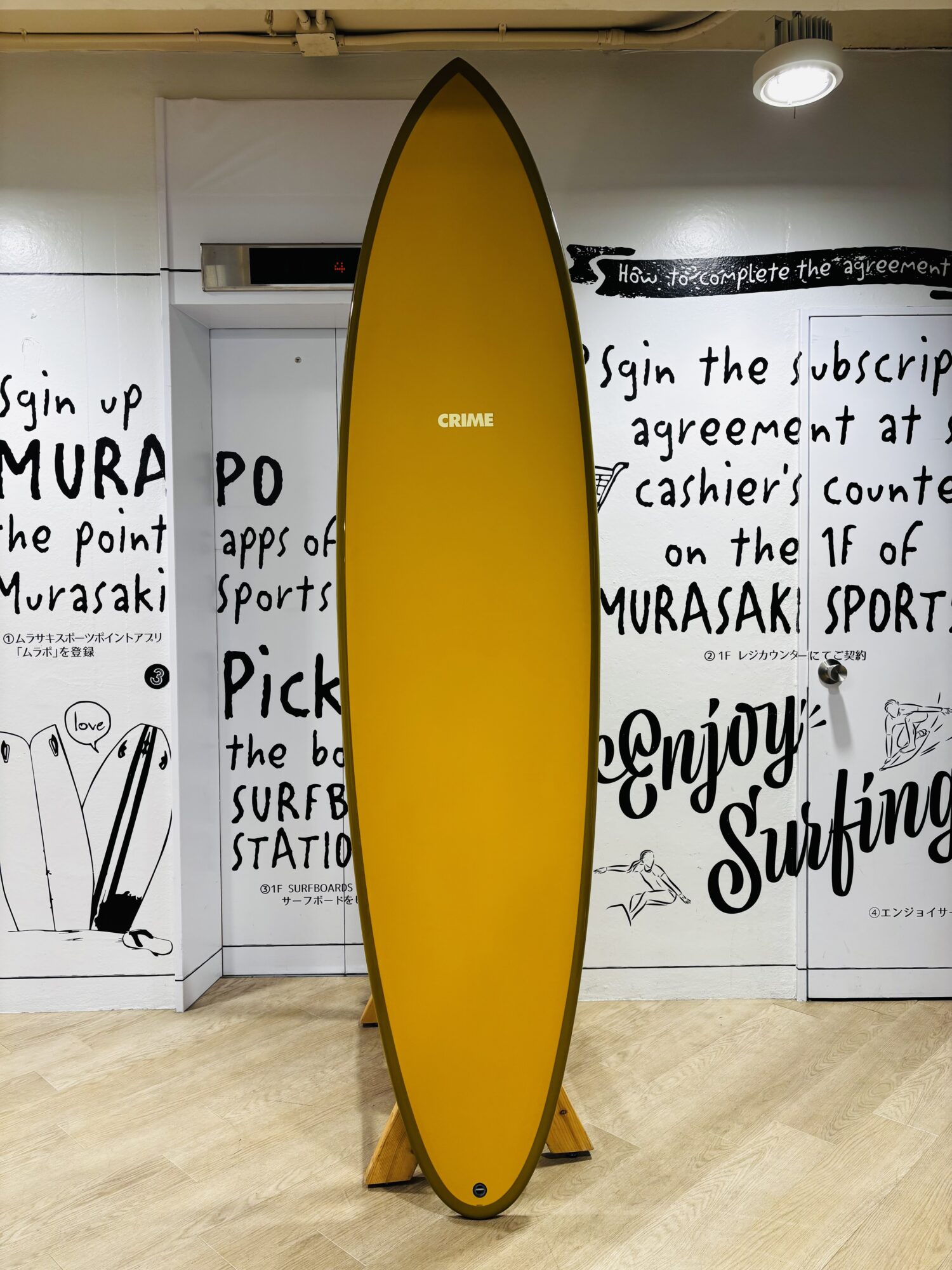 Yellow surfboard standing upright on a wooden floor indoors, with black graffiti-style text on white walls behind it and 'CRIME' written on the board center.